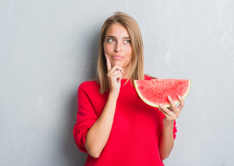 Beautiful young woman over grunge grey wall eating water melon serious face thinking about...