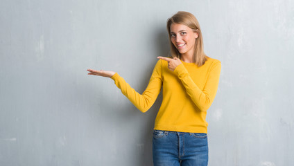 Beautiful young woman standing over grunge grey wall amazed and smiling to the camera while presenting with hand and pointing with finger.