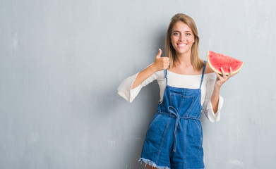 Beautiful young woman over grunge grey wall eating water melon happy with big smile doing ok sign, thumb up with fingers, excellent sign