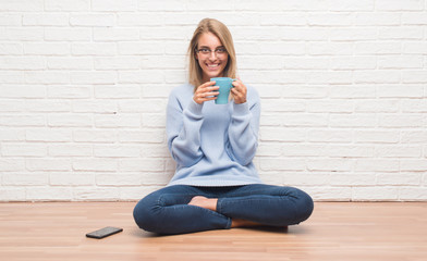 Fototapeta na wymiar Beautiful young woman sitting on the floor driking cup of coffee at home with a happy face standing and smiling with a confident smile showing teeth