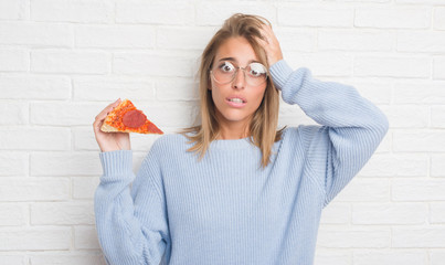 Beautiful young woman over white brick wall eating pizza slice stressed with hand on head, shocked...