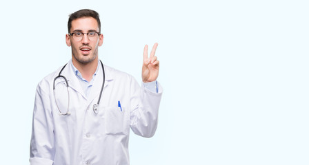 Handsome young doctor man smiling with happy face winking at the camera doing victory sign. Number two.