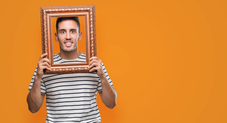 Handsome young man looking through vintage art frame with a happy face standing and smiling with a confident smile showing teeth