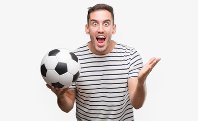 Handsome young man holding soccer football very happy and excited, winner expression celebrating victory screaming with big smile and raised hands