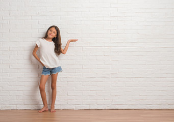 Young hispanic kid stading over white brick wall smiling cheerful presenting and pointing with palm...