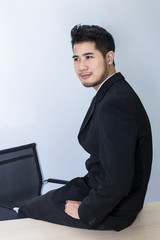 Young handsome businessman smiling and smart sit on table at office.He is 20-30 years old in black suit.Copy space.