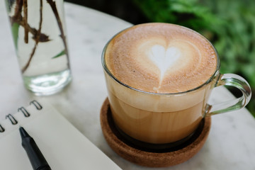 Close up caffe latte coffee in a glass cup on a marble coffee table with white notebook black pen glass vase with green background