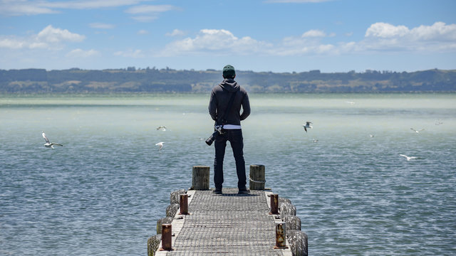 Young Asian male photographer standing on the jetty looking at scenery of lake in Rotorua, North Island, New Zealand. Travel and photography concepts