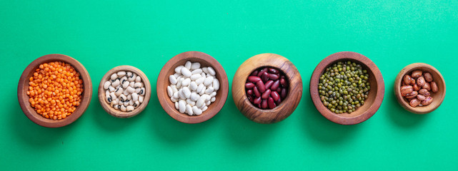 Assortment of legumes pulses in wooden bowls on green background, isolated, top view, banner.