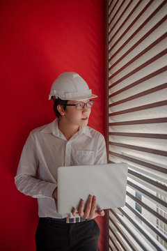 Young Asian Male Engineer Or Architect Wearing Protective Helmet And Glasses Working With Laptop Computer With Red Wall In The Background. Architecture, Engineering And Building Construction Concepts