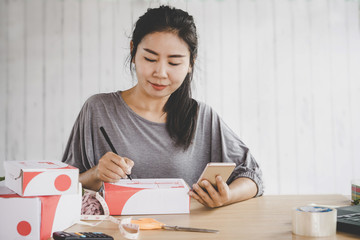 beautiful Asian woman selling online from home writing on box preparing order with smart phone in hand 