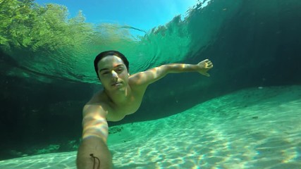 POV underwater view of young man swimming in crystal clear tropical blue water on sunny day with lens flare