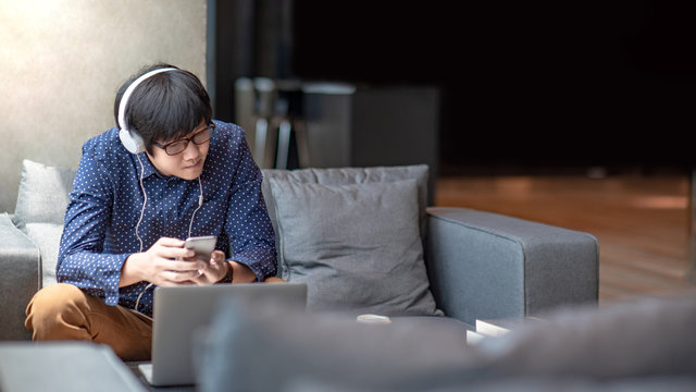 Young Asian man using smartphone for listening to music on mobile application, sitting and relaxing on sofa during free time. Urban lifestyle in living space concept