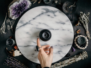 Witch's hand specifies on the black candle on a marble white round tray. The place for witchcraft with magic things around. View from above - obrazy, fototapety, plakaty