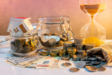 Saving money for retirement concept. Real estate or property investment. Home mortgage loan rate. Coin stack, money bag and currency glass jars on banknotes with hourglasses, house model on the table.