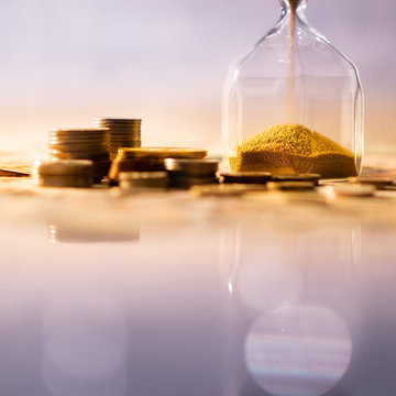 Reflection of brown sand running through the shape of modern hourglass on glowing table with currency. Time investment and passing time. Urgency countdown timer for business deadline concept