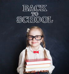 Cute little girl pupil holding pile of books and smiling. Schoolgirl in glasses on blackboard...