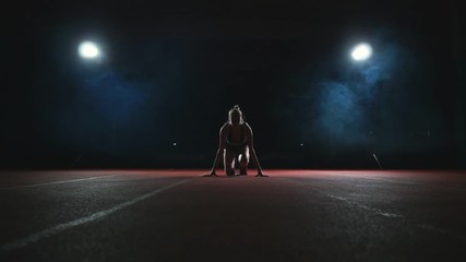 Slender young girl athlete is in position to start running in the pads on the track in slow motion against the smoke - Powered by Adobe