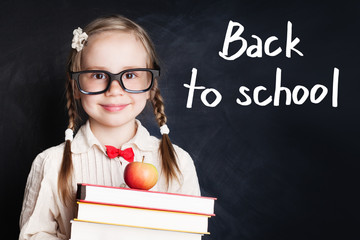 Smiling child girl in school uniform holding books and apple against chalk board background. Back...