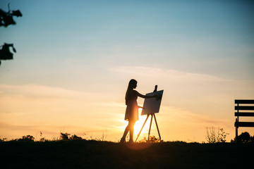 Naklejka premium Silhouette of a blonde girl artist. Lady paints a painting on the canvas with the help of paints. A wooden easel keeps the picture. Summer is a sunny day, sunset