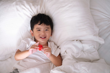 Obraz na płótnie Canvas 3 years old little cute Asian boy at home on the bed, kid lying playing and smiling on white bed with pillow and blanket, top view with copy space.