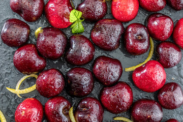 Precious red and fresh cherry pints, fresh and raw on a textured and veined black background. With lemon yellow zest and peppermint mint leaves.