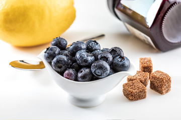 Fresh and raw blueberries in delicate porcelain bowl on white napkin and spoon. Includes brown sugar and lemon. On white background.