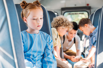 depressed little schoolgirl sitting alone in school bus while her classmates using smartphone together on background