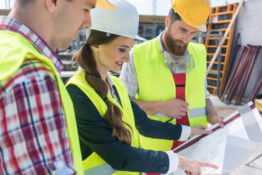 Young and confident female architect smiling while sharing ideas about the plan of a building under construction outdoors with two blue-collar workers