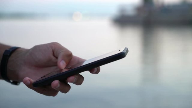 Closeup of a male hand keeping a black phone and touching its screen while seeking some information on a river quay at picturesque sunset