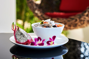 Concept of colorful natural products arranged in white bowl for breakfast. Mix of pitaya and exotic fresh fruits lying in plate with violet petals for healthy lunch.