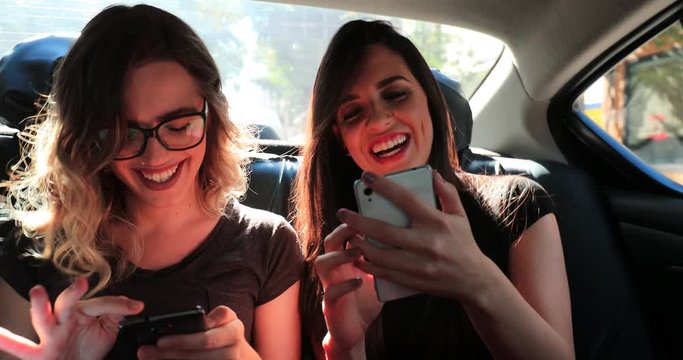 Female friends browsing on their smartphone while riding cab laughing. Women laughing while using cellphone in the back seat of a car2