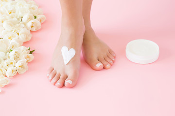 Woman's perfect, groomed feet with jar of natural herbal cream. Care about clean, soft and smooth skin. Heart shape created from cream. Love a body. Beautiful roses on pink background. Fresh flowers.