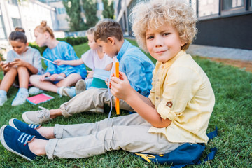 confused curly schoolboy holding book while sitting on grass with classmates