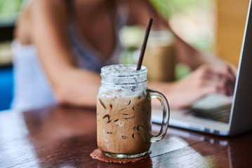 Close-up glass coffee with blur women working on laptop computer in background.