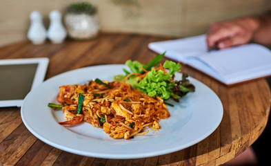 Healthy business lunch in the cafe, view of dish on wooden table near tablet. Thai stir-fried rice noodles. Snack at break time.