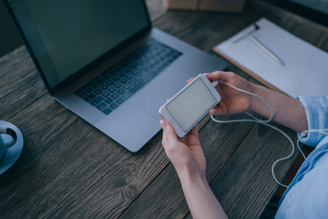 Female hands holding contemporary smart phone while sitting at desktop with laptop computer. Business woman updating applications on cellular. Mockup image of a woman's hand holding mobile phone.
