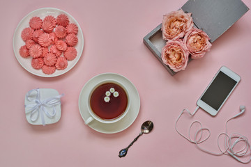 Obraz na płótnie Canvas A cup of hot herbal tea, flowers and a smartphone on pink table. Pink peony-shaped roses in gift box and marshmallow on the plate. Female desktop. Workplace for creativity plans and dreams. Top view.
