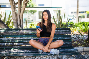 Pretty latin girl using a mobile phone sitting in a bench in an urban park