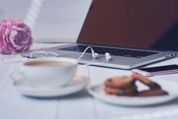 Hot cup of fresh coffee and laptop on the work desk. Cosy home and work concept - vintage wooden table with laptop computer, coffee cup and biscuit. Women's elegant desktop. Selective focus.