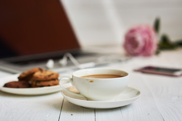 Hot cup of fresh coffee and laptop on the work desk. Cosy home and work concept - vintage wooden table with laptop computer, coffee cup and biscuit. Women's elegant desktop. Selective focus.