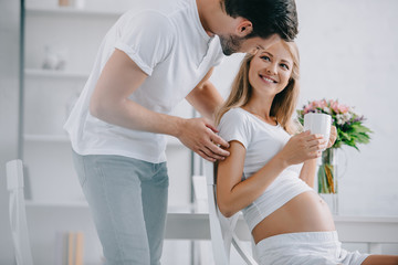 smiling pregnant woman with cup of tea resting on chair with husband near by at home