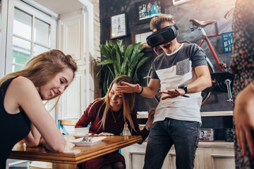 Young women laughing at their emotional male friend playing horror game in virtual reality goggles...