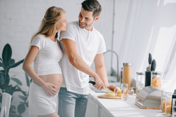 portrait of smiling man cutting fruits at counter with pregnant wife near by in kitchen at home
