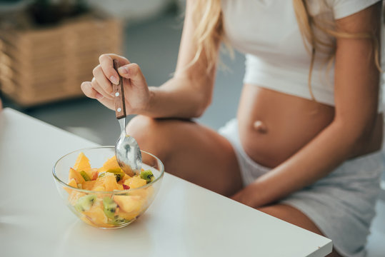 partial view of pregnant woman with spoon and fruits salad in bowl at home