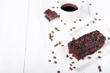 Black tea in a white cup with chocolate, chocolate cake on a white saucer and coffee beans on a white wooden background