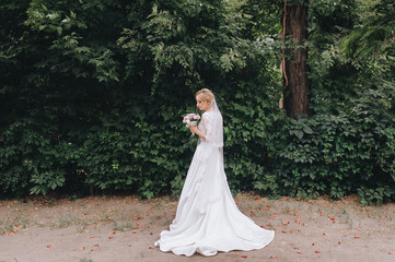 A beautiful bride in a lace dress is standing with a bouquet in her hands against the backdrop of...