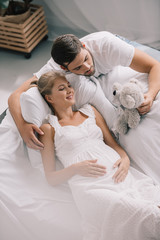 Obraz na płótnie Canvas high angle view of man with teddy bear near pregnant wife in white nightie on sofa at home