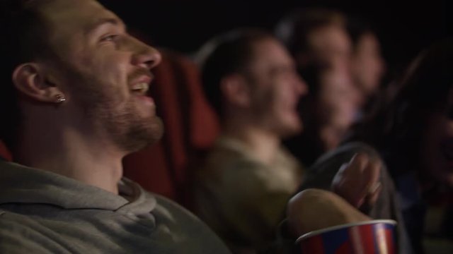 Young guy eating popcorn in slow motion. Young man laughing in movies theater. Hipster man eating popcorn in cinema. Close up of man watching comedy film