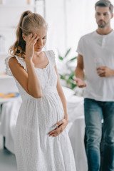 pregnant woman in white nightie having headache while husband with medicines and glass of water standing behind at home